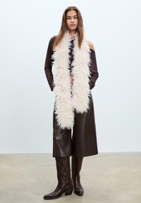 Lunga sciarpa in crema, morbida e realizzata in pelliccia ecologica, indossata sopra un top a maniche lunghe scuro e culottes in pelle marrone, completata da stivali da cowboy marroni.