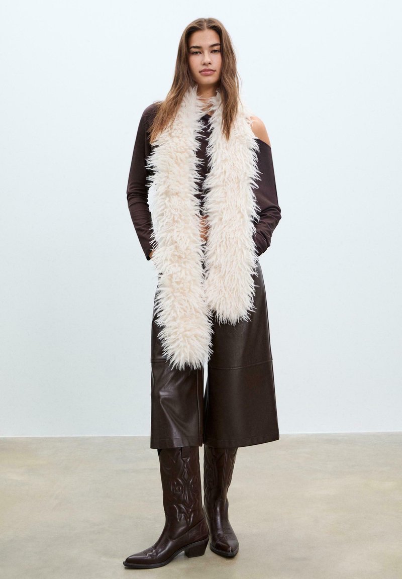 Lunga sciarpa in crema, morbida e realizzata in pelliccia ecologica, indossata sopra un top a maniche lunghe scuro e culottes in pelle marrone, completata da stivali da cowboy marroni.