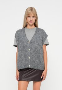 Gray knitted vest with a v-neckline, short sleeves, and five buttons down the front, worn over a gray t-shirt and a dark leather skirt.