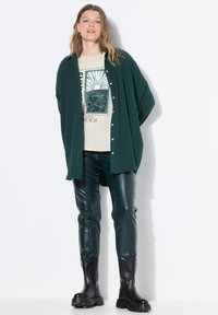 Camicia verde con bottoni, t-shirt graphic color crema, pantaloni in faux pelle verde e stivali neri con suola chunky, in piedi contro uno sfondo bianco.