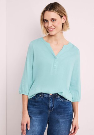 Vrouw met kort blond haar, gekleed in een lichtblauw drieknoopsblouse en een blauwe spijkerbroek, glimlachend tegen een effen lichte achtergrond.