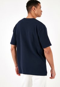 T-shirt din bumbac de culoare albastru marin, cu mâneci scurte rulate, croială lejeră și textură subtilă, văzut din spate pe un fundal alb.