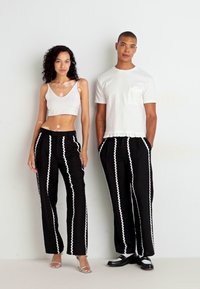 Pantalones negros con un patrón de zigzag blanco, combinados con un top corto blanco ajustado y una camisa blanca de manga corta, delineada con material texturizado.