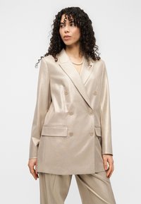 Διπλόμπουτο σακάκι από γυαλιστερό μπεζ ύφασμα με δύο εμπρόσθιες τσέπες και κυκλικά κουμπιά, σε συνδυασμό με φαρδιά μπεζ παντελόνια.