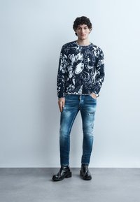 Mladý muž stojí opretý o sivú stenu, oblečený v námornícky modrom svetri s kvetinovým vzorom, roztrhaných úzkych džínsoch a čiernych kožených topánkach.