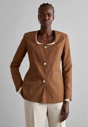 Blazer taillé marron avec un col rond, fermeture à un bouton et deux poches avant, porté sur un pantalon crème texturé.