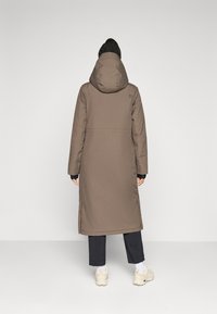 Persona con capelli intrecciati, che indossa un lungo cappotto marrone con cappuccio, pantaloni neri, calze bianche e sneakers beige, di spalle su uno sfondo neutro.