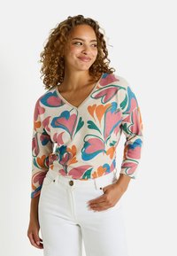 Un pull à motifs floraux multicolores avec un col en V, des manches trois-quarts et un fond beige, associé à un jean taille haute blanc.