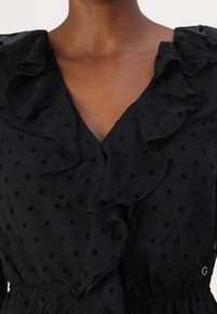 Vestido negro con textura de lunares, escote con volantes, forma en V y cintura elástica. El material transparente añade suavidad y dimensión.