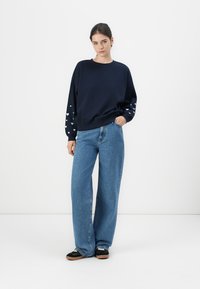 Mørkeblå sweatshirt med hvitt hjerte mønster på ermene, kombinert med lyseblå vide jeans. Enkelt design, myk stoff, avslappet passform.