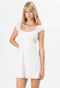Kvinna med långt blont hår som bär en kort vit klänning med rosa blommönster och puffärmar, stående mot en enkel vit bakgrund.