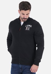 Sweatshirt zippé noir en tissu doux, avec un col haut, des poches latérales et un logo imprimé blanc sur la poitrine.