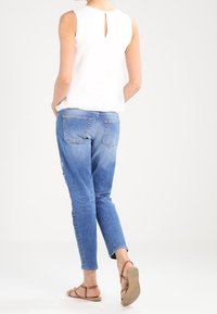 Top bianco senza maniche con dettaglio a chiave sulla schiena, abbinato a jeans slim-fit blu chiaro e sandali piatti color beige.