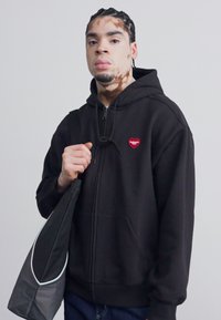 Juodas zip-up gobtuvėlis su raudona širdies logo ant krūtinės, derinamas su pilku krepšiu. Lygus audinys, laisvas kirpimas ir reguliuojamas gobtuvas.