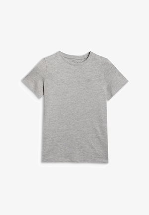 Jeune fille debout, portant un t-shirt gris à manches courtes sur un t-shirt blanc à manches longues, un pantalon rose clair, des baskets blanches et un bandeau blanc.