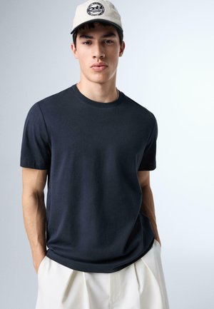 Mladý muž oblečený v čiernom tričku s okrúhlym výstrihom, bielych nohaviciach a bielej šiltovke s čiernym logom, stojaci s rukami v kapse.