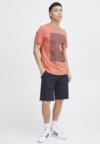 Korallfärgad t-shirt med svart randig grafik av en palm, ihop med navy shorts. Vita idrottsskor med svarta detaljer.