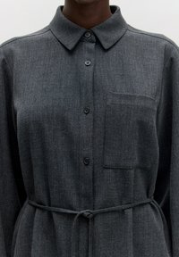 Grijze button-up overhemd gemaakt van textuurstof. Bevat een kraag, borstzak en een vastgebonden tailleband. Knopen zijn donker en rond.