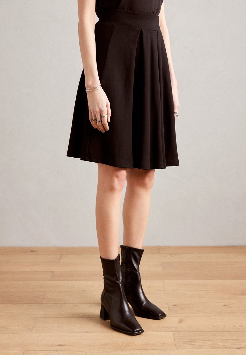 Femme portant une jupe noire longueur genoux et des bottines noires à talons, debout sur un sol en bois face à un mur uni.