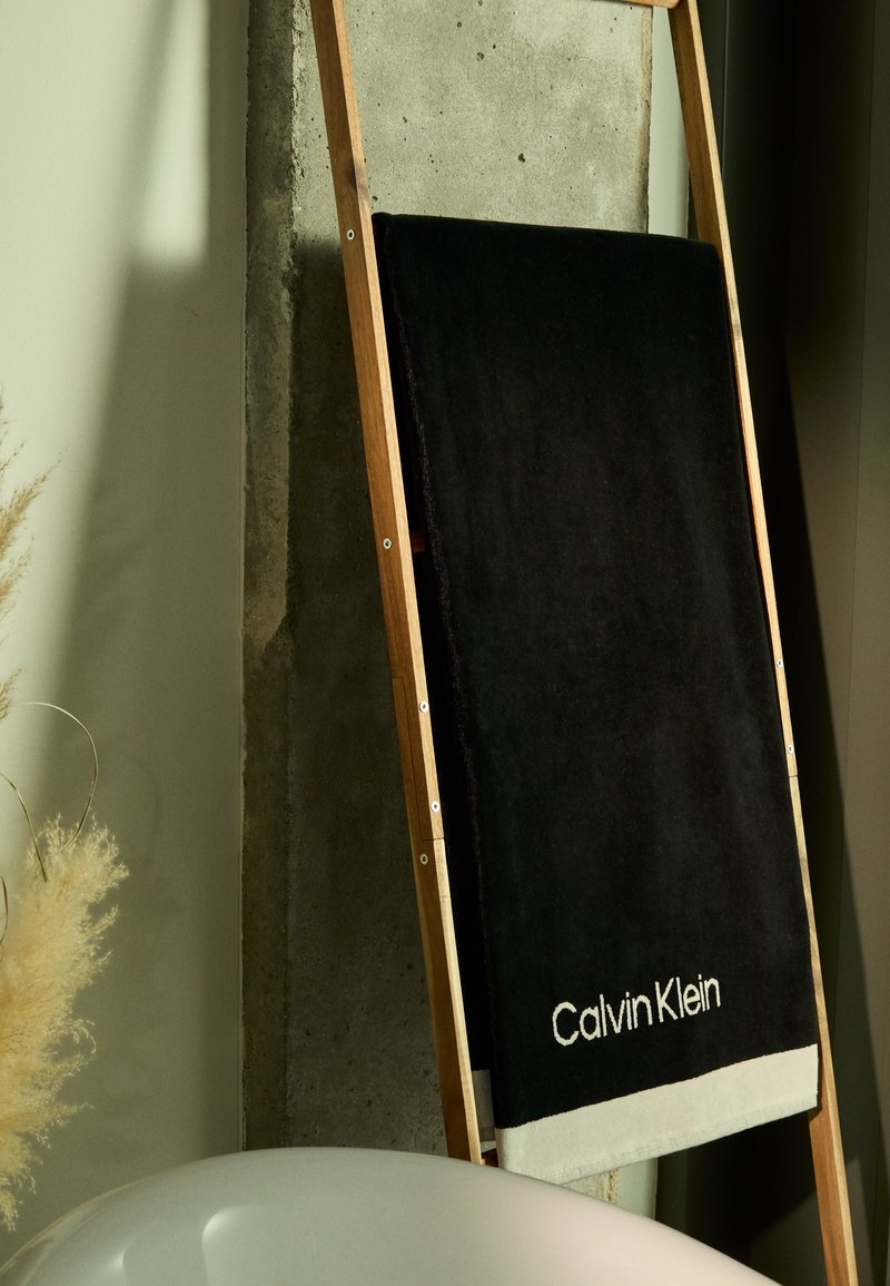 Zwart handdoek met beige rand, waarop het "Calvin Klein" logo staat, hangend aan een houten ladder tegen een betonnen muur. Zachte textuur en minimaal ontwerp.