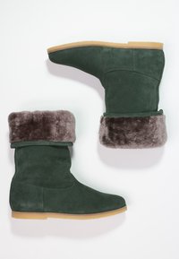 Bottes en daim vert foncé avec un doux revers en fourrure grise, bout arrondi et semelles en caoutchouc. Le design présente une silhouette simple et épurée.