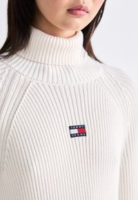 Suéter de punto blanco acanalado con un cuello alto. Presenta un pequeño parche de tela con un logo en rojo, blanco y azul con el texto "TOMMY JEANS".
