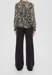 Blusa nera trasparente con motivi astratti bianchi e beige, con polsini arricciati, abbinata a jeans neri larghi. Tessuto testurizzato con una vestibilità ampia.