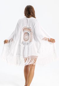 Kaftano bianco con orlo frangiato e dettagli a crochet sulla schiena. Tessuto leggero, design aperto con maniche larghe e motivo circolare.