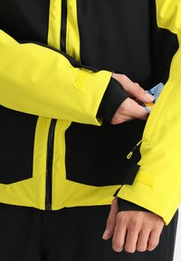 Veste imperméable jaune et noire avec fermeture éclair, détail de logo en tissu et accès à une poche sur la manche pour des cartes ou de petits objets.