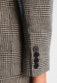 Brun och beige hounds-tooth blazerärm med fyra svarta knappar och texturerat tyg. Skräddarsydd passform med strukturerad design.