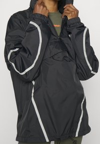 Chaqueta cortavientos negra con acentos reflectantes blancos, material ligero, corte oversize, capucha ajustable y puños elásticos.