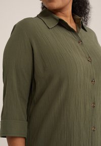 Camicia button-up verde oliva con texture e colletto, dotata di bottoni in legno, maniche arrotolate e vestibilità casual.