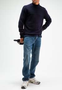 Marineblauwe trui met geribbelde structuur en hoge kraag, gecombineerd met lichtblauwe jeans en witte sneakers met accenten. Houdt een zwarte hoed vast.