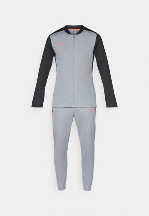 Ensemble de survêtement Nike gris et noir avec une veste à zip intégral et un pantalon fuselé, arborant des logos swoosh orange sur la poitrine et la cuisse.