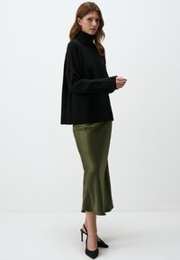 Zwarte oversized coltrui, zachte textuur, geribbelde manchetten. Olijfgroene satijnen midi-rok, gladde afwerking, uitlopende zoom. Zwarte puntige hakken.