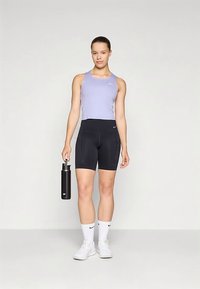 Jauna sieviete sportiskā apģērbā tur melnu ūdens pudeli, ģērbta gaiši violetā tanku topā, melnās šortos, baltās Nike zeķēs un baltos treniņbiksēs.