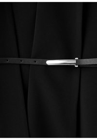 Curea subțire din piele neagră cu o cataramă dreptunghiulară argintie lucioasă, purtată peste o ținută din material negru.