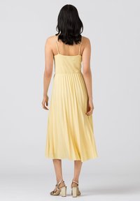 Gul plisserad klänning med tunna axelband, medellängd, lättviktsmaterial, flödande design, bärs med metalliska klacksandaler.