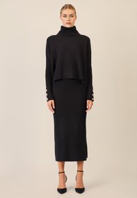 Zwarte gebreide turtleneck trui met een cropped ontwerp, gecombineerd met een lange, geribbelde zwarte rok met een zijspleet en knoopaccenten op de manchetten.