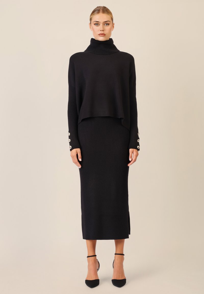 Zwarte gebreide turtleneck trui met een cropped ontwerp, gecombineerd met een lange, geribbelde zwarte rok met een zijspleet en knoopaccenten op de manchetten.
