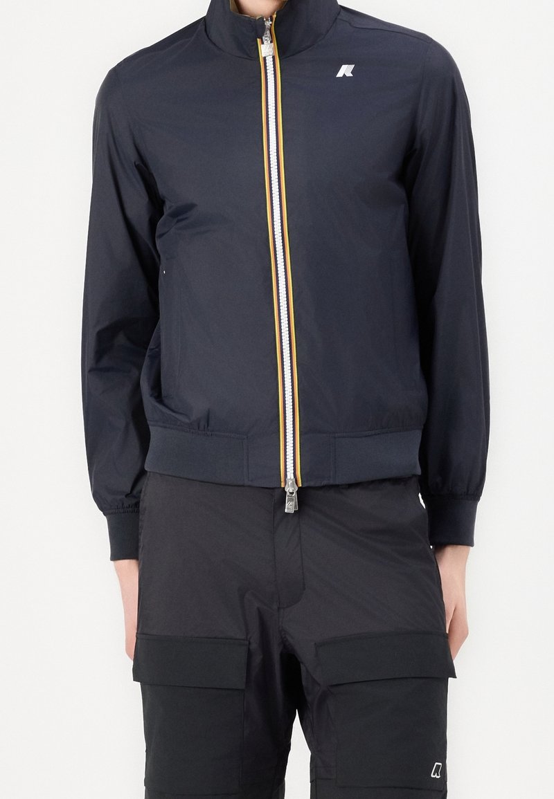 Homme portant une veste zippée bleu marine avec un petit logo blanc et un pantalon cargo noir avec des poches à rabat à l'avant.