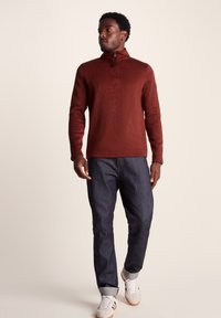 Brown zip-up pullover in a textured knit, paired with dark denim jeans and light-colored sneakers. Simple, modern design without logos.