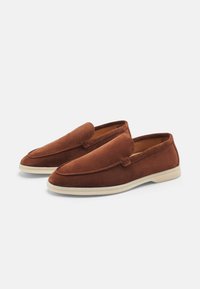 Mocassins en daim marron avec une texture lisse, un bout arrondi et un design à enfiler. Présente des coutures minimalistes et une semelle extérieure blanche contrastante.