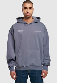 Grå hettegenser i mykt stoff, med frontlomme, ribbede mansjetter og to logoer på brystet. Båret sammen med blå jeans.