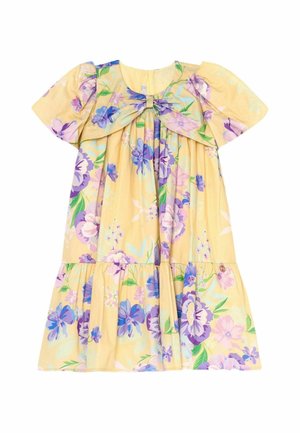 Ung pige med krøllet hår iført en gul kjole med lilla blomster og beige flettede remsandaler, gående mod en ensfarvet beige baggrund.