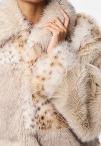 Beżowy płaszcz z futerka ekologicznego o miękkiej, puszystej fakturze z cętkami przypominającymi lamparta. Kołnierz i mankiety są grube i obfite.