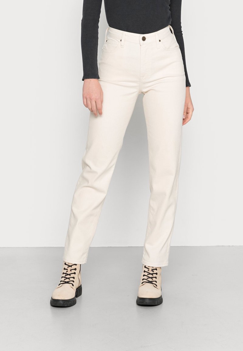 Personne portant un pantalon blanc droit, un haut noir à manches longues et des bottes beiges à lacets debout sur un sol gris clair.