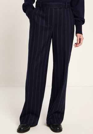 Person wearing dark navy wide-leg trousers with vertical stripes, paired with black shoes and a dark sweater, standing with hands near pockets.