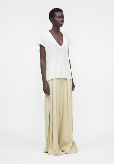 Woman with short curly hair wearing a white V-neck short-sleeve top and wide beige pleated pants standing against a plain background.