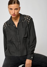 Chemise noire avec des accents de œillets argentés sur les épaules, deux poches avant, design à boutons, et manches longues avec des poignets à bouton.
